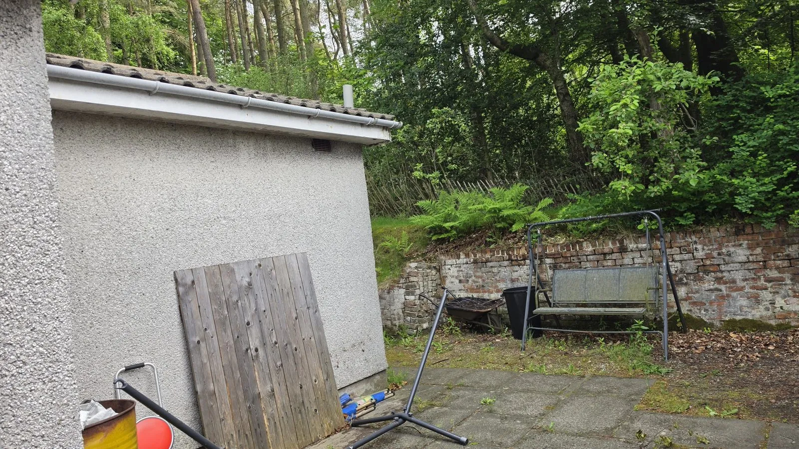 Old concrete slabs beside a pebbledash wall, with a metal swing, a brick wall and trees behind.