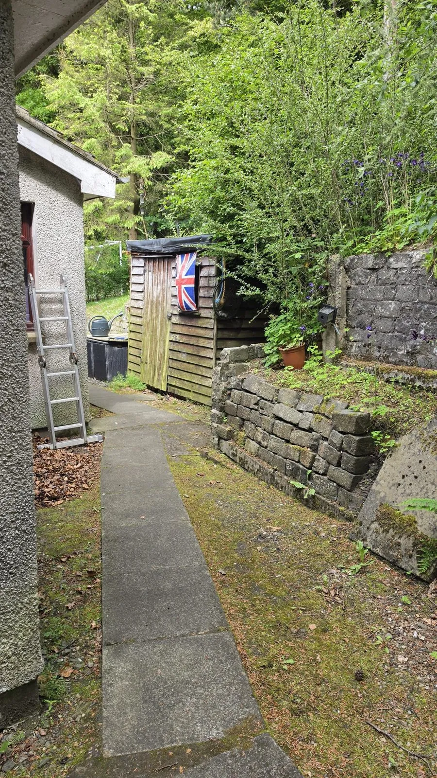 A narrow concrete path between a pebbledash wall and an old retaining wall, with a timber shed.