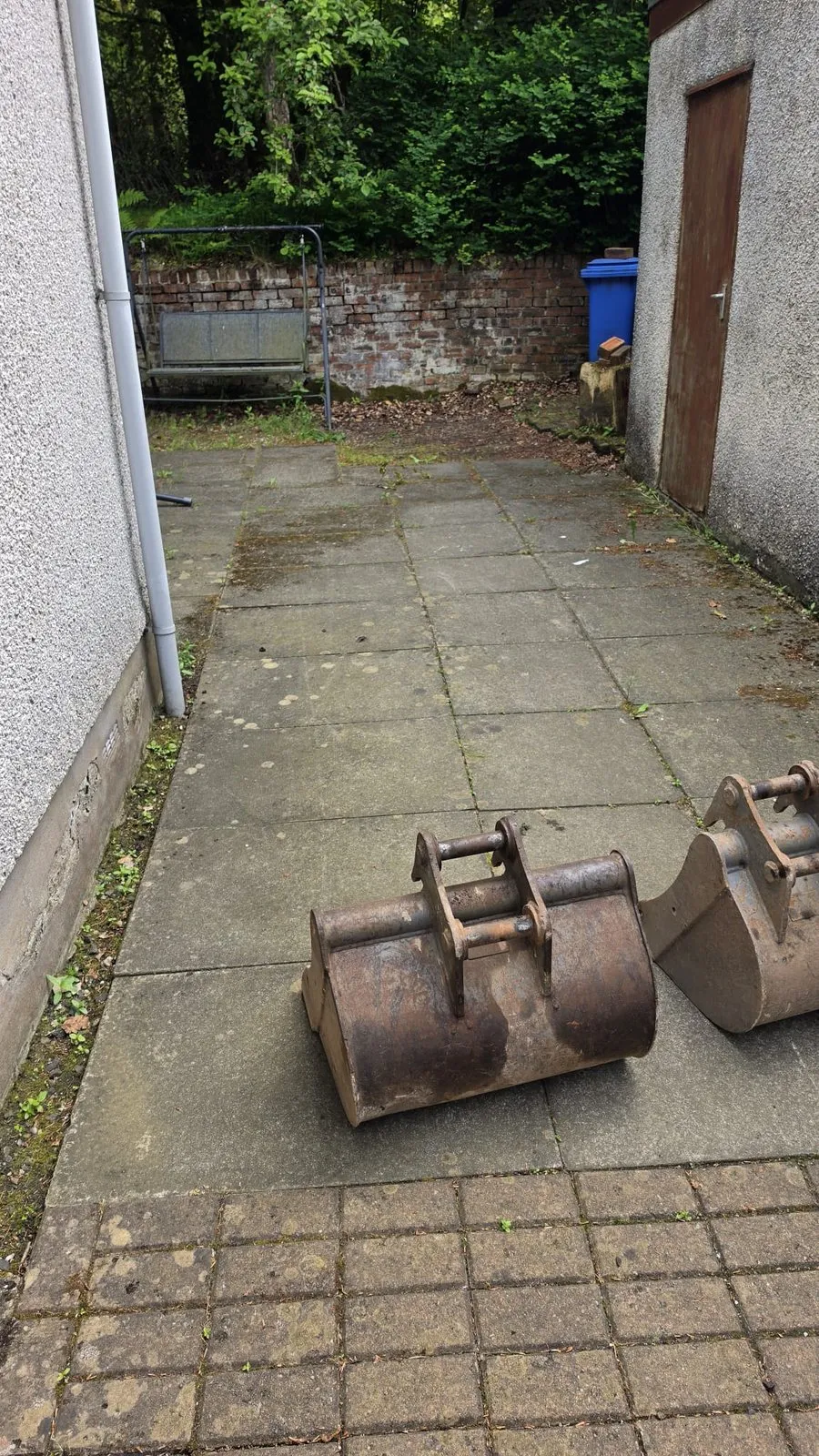 A run of stained concrete slabs between two rendered walls, with excavator buckets in the foreground.