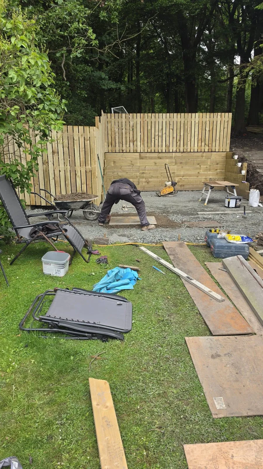 A gravel base in front of a new timber fence, with boards and tools on the grass and a person setting a slab.
