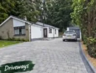Grey block-paved driveway beside a white house, edged and running up to a garage with a silver car.