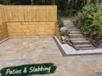 Mixed grey and buff paving slabs beside a timber fence, with steps rising through a planted bank.