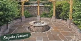 Circular stone fire pit under a timber pergola, laid in radial sandstone with a low stone wall and shrubs.