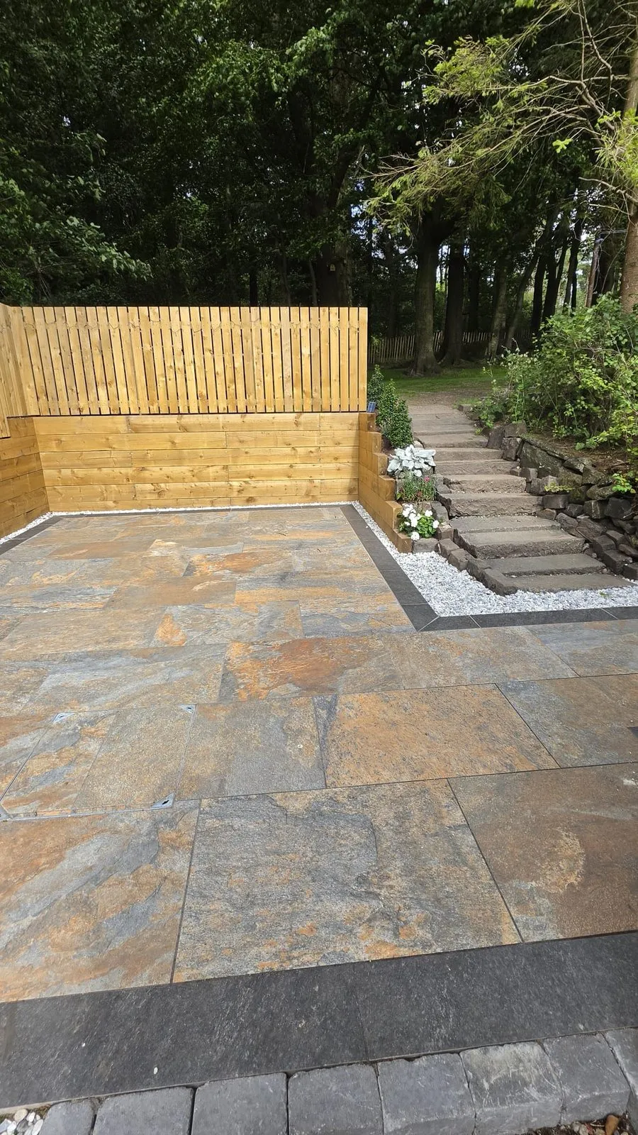 A finished mixed-stone patio edged in dark slate, with a timber fence and stone steps up a wooded bank.