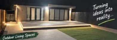Night photograph of a lit timber deck outside bifold doors, with pale paving and a lawn in front.