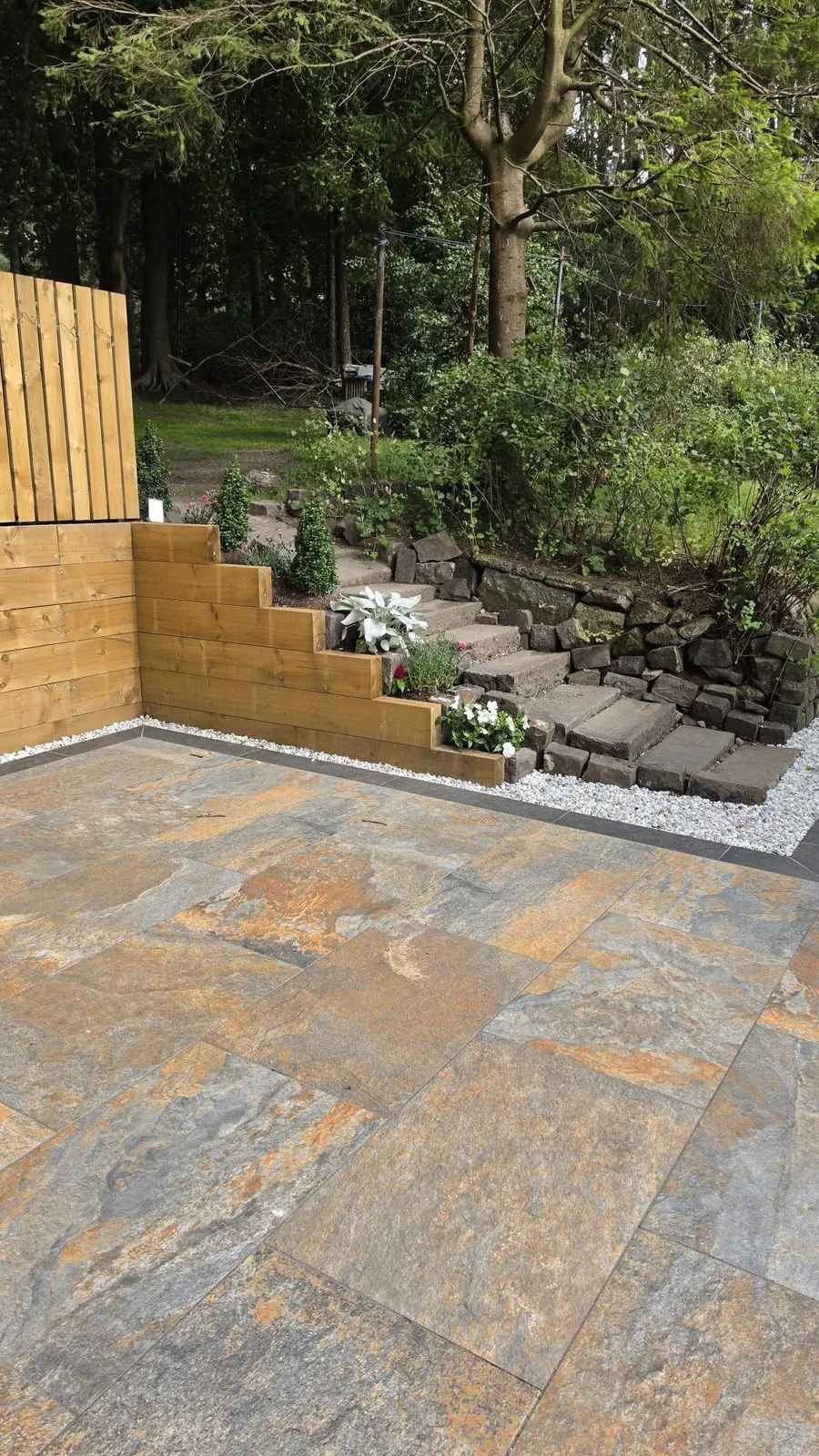 Close view of mixed stone paving, a timber planter of white flowers, and stone steps into the trees.