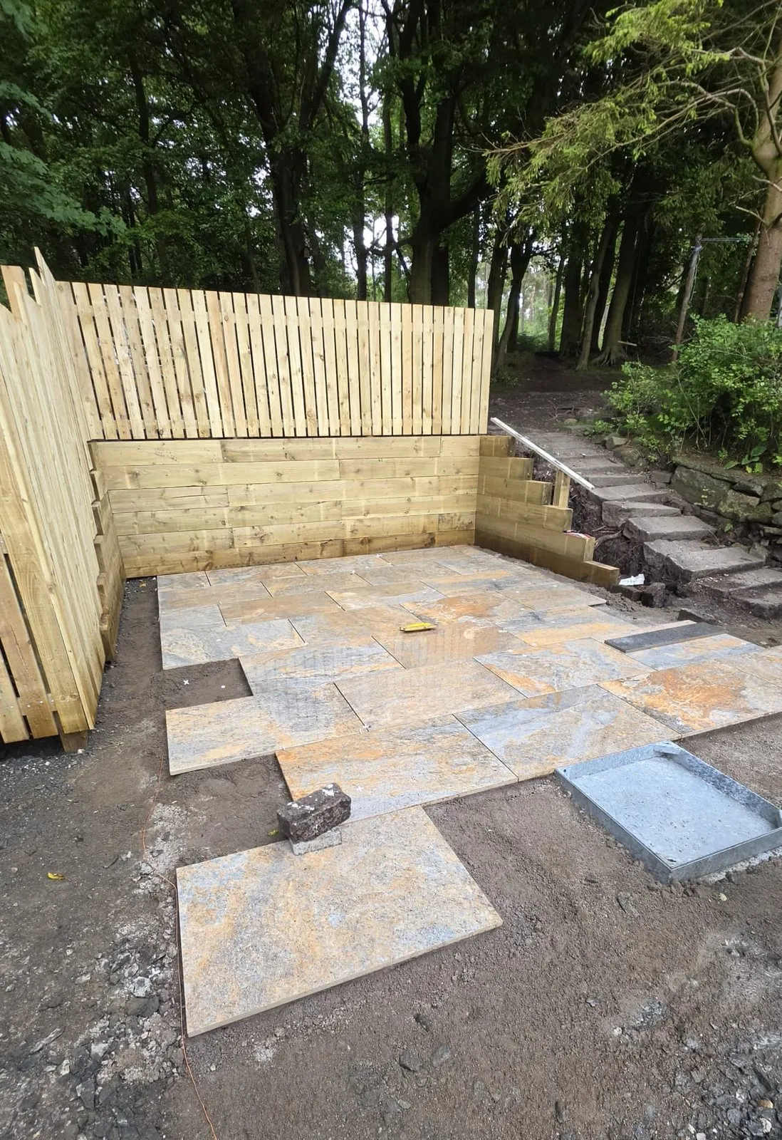 Mixed stone slabs part-laid inside a new timber fence, with loose slabs and a manhole cover still on the soil.